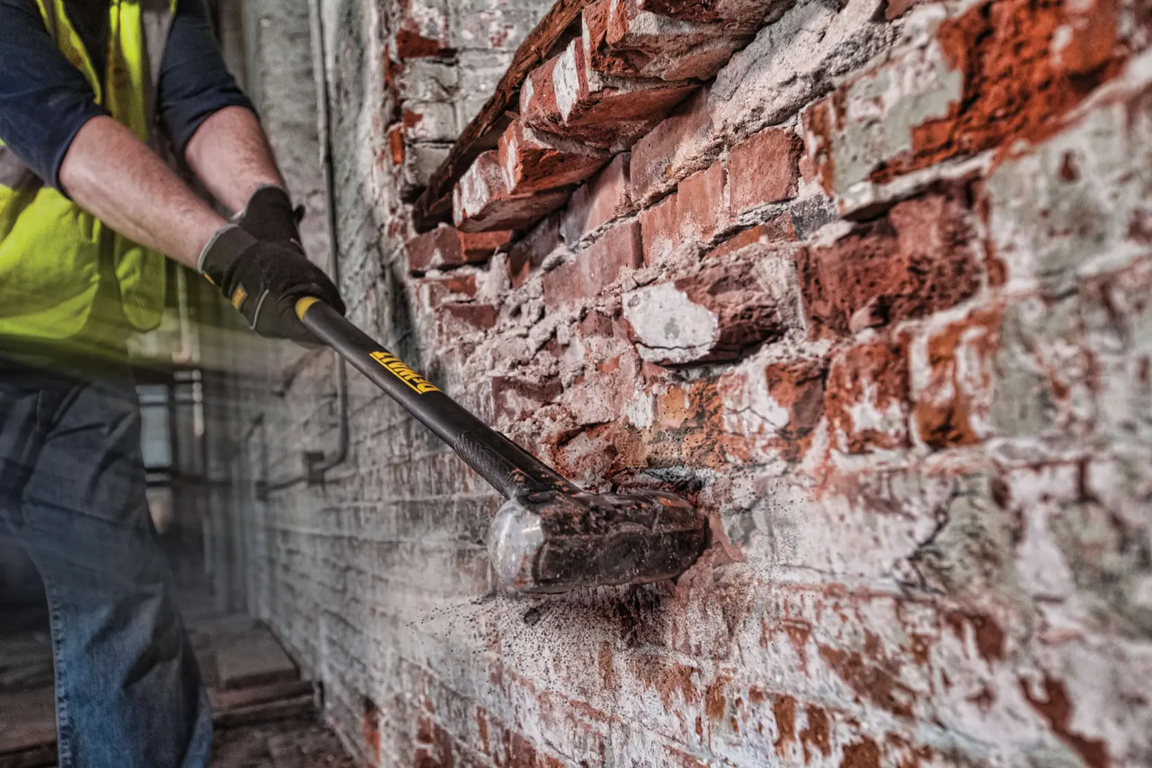 Una persona con guanti e giubbotto di sicurezza usa una mazza DEWALT (modello DWHT56029) per rompere un muro di mattoni. Polvere e detriti si disperdono quando la mazza colpisce i mattoni.