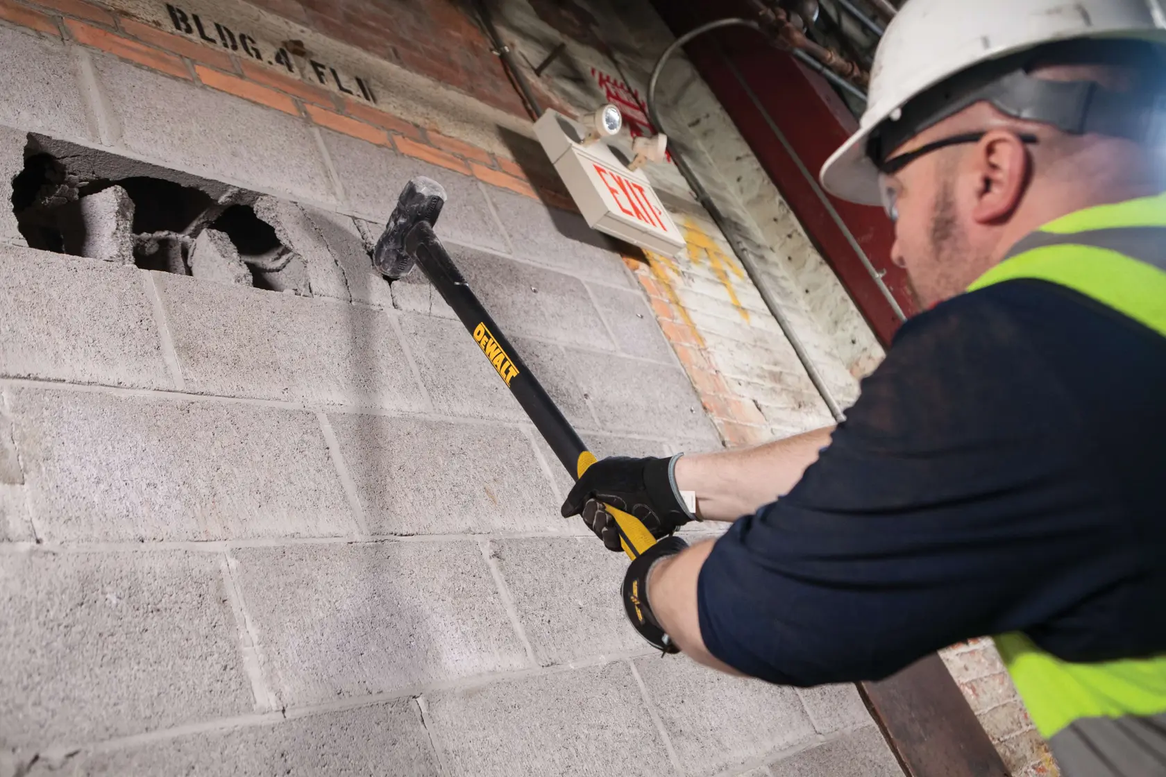 Una persona con equipaggiamento di sicurezza utilizza una mazza DEWALT (modello DWHT56029) per demolire una parete in blocchi di cemento all'interno.