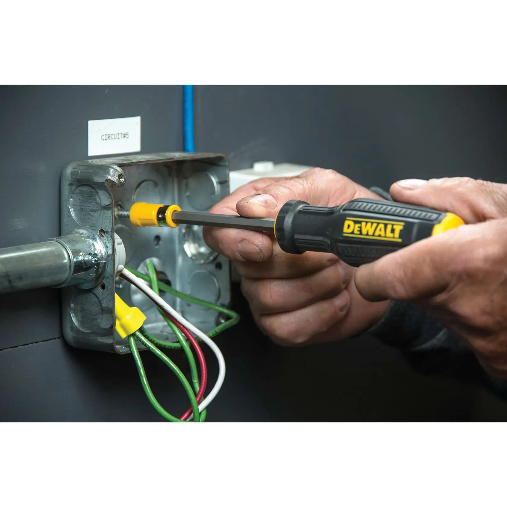 A person using a DEWALT screwdriver to tighten a screw inside an electrical junction box, with visible colored wires.