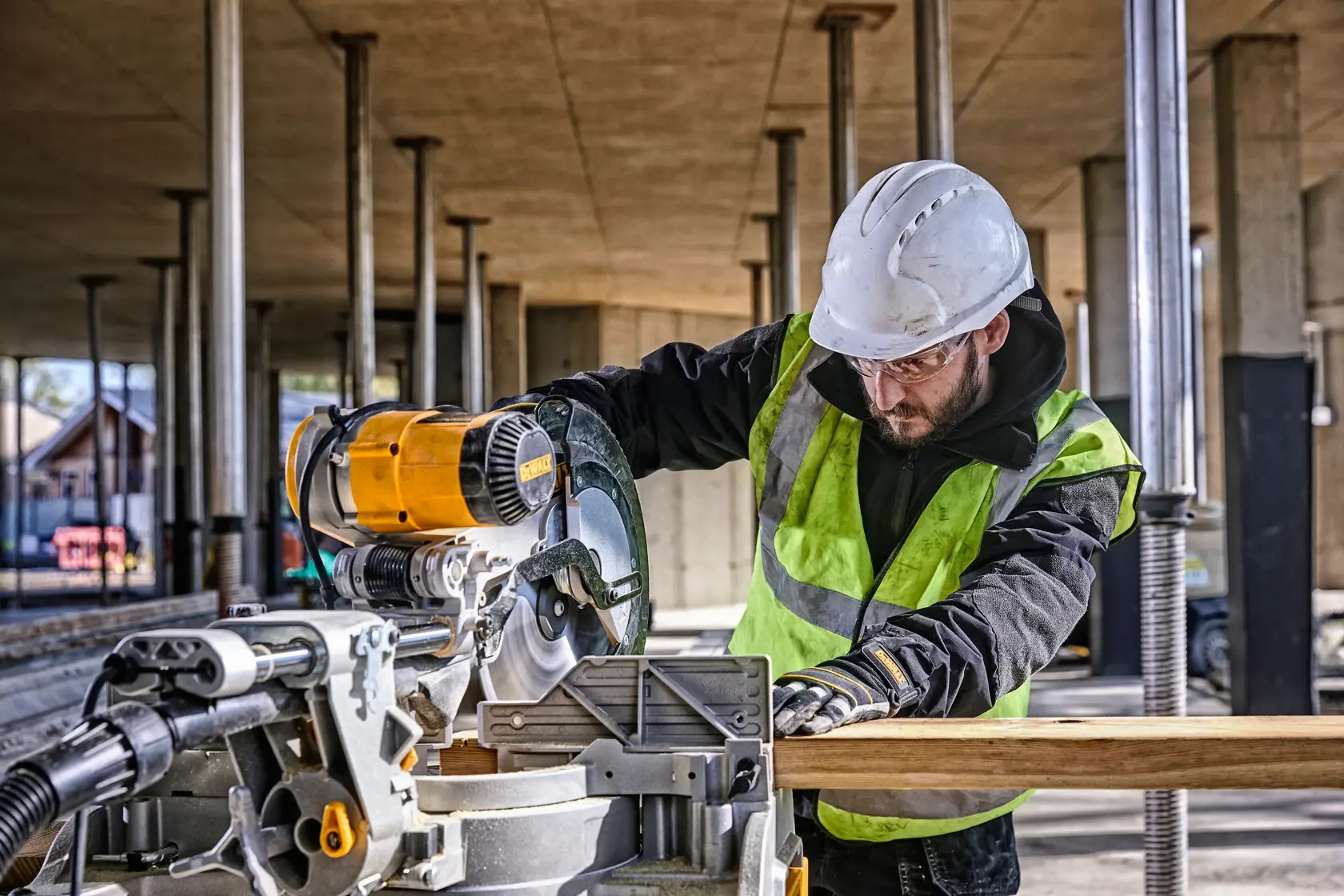 A DEWALT DWS780 miter saw being used to cut a wooden plank at a construction site. The operator is wearing a safety helmet, gloves, and a high-visibility vest. The background shows a partially constructed building with vertical pillars.