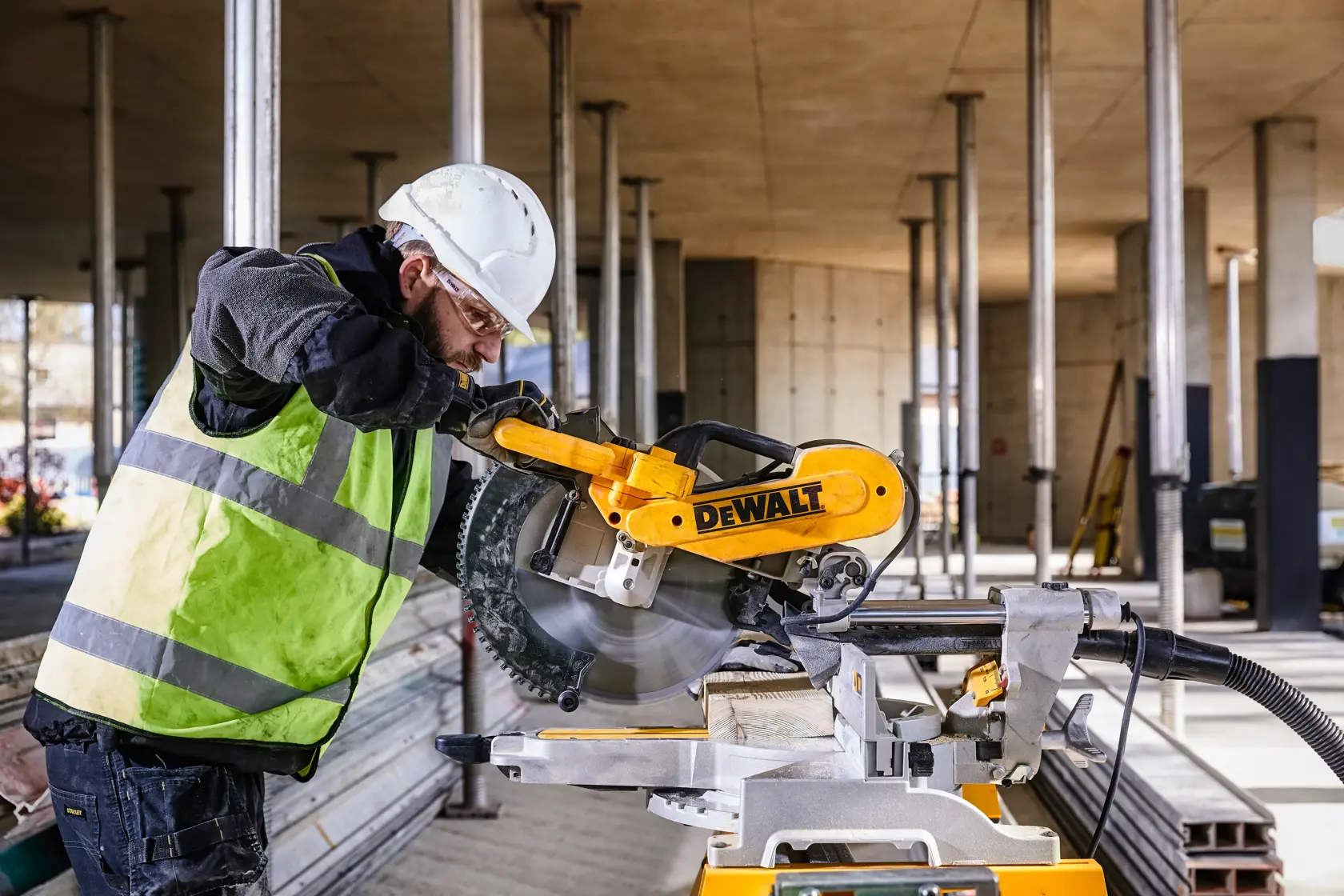 A person wearing a white safety helmet and gloves operates a yellow DEWALT DWS780 miter saw at a construction site. The background shows an unfinished building structure with columns and beams.