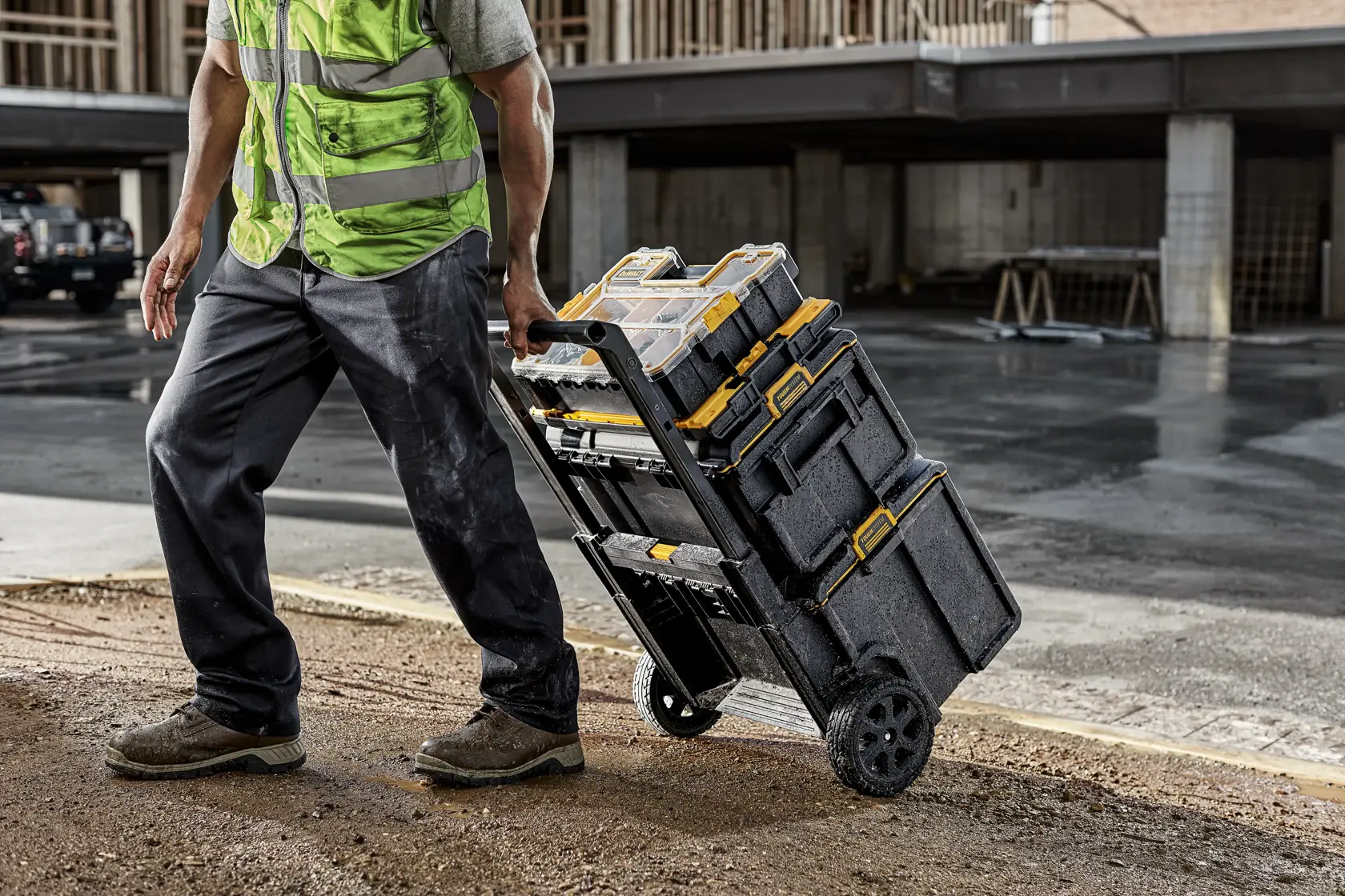 Une personne portant un gilet haute visibilité utilise un chariot à roulettes pour transporter des boîtes à outils empilées DEWALT sur un chantier.