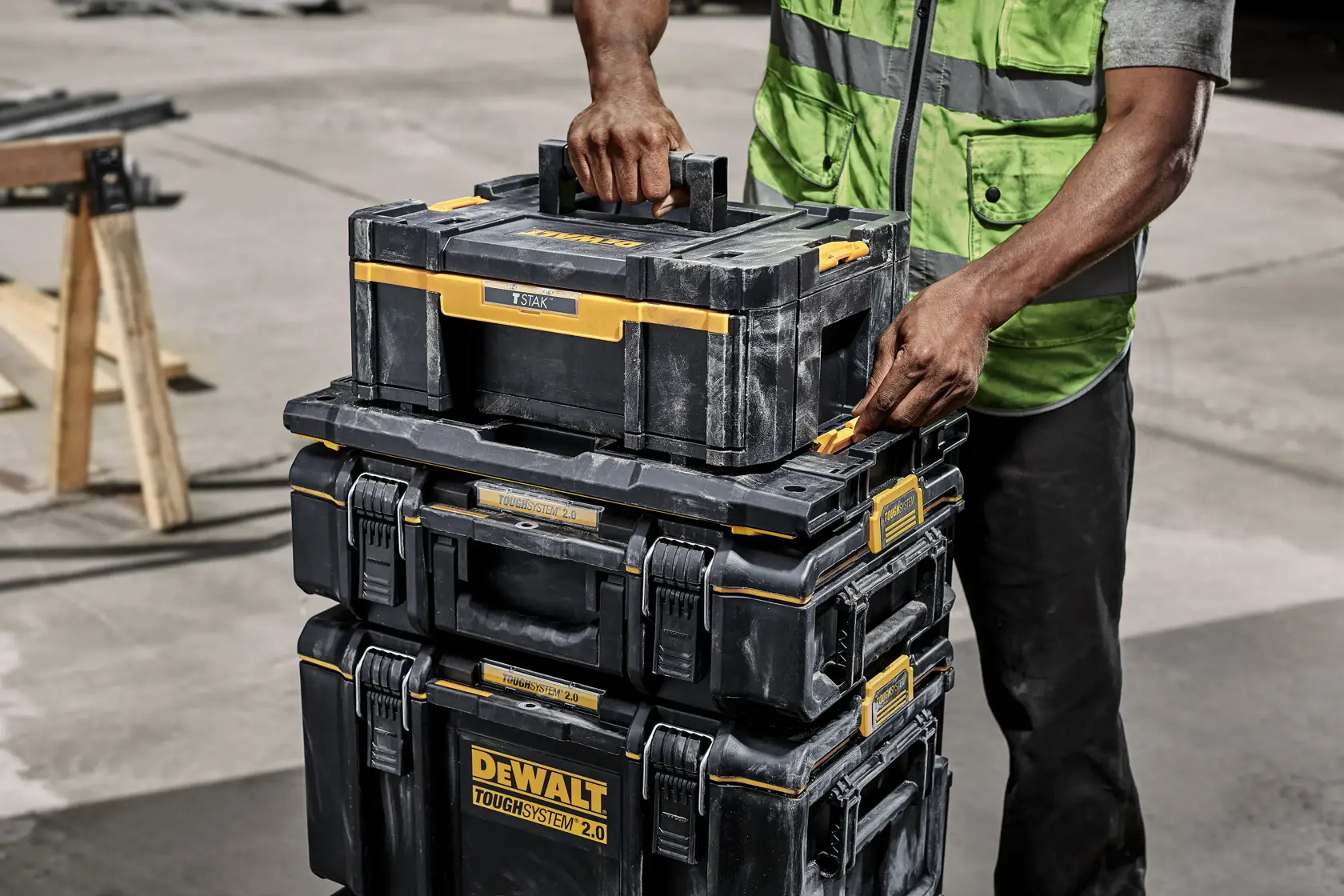 Une personne portant un gilet de sécurité vert manipule une pile de boîtes à outils noires et jaunes DEWALT ToughSystem 2.0 à l'extérieur, en mettant l'accent sur la boîte supérieure qui est soulevée.