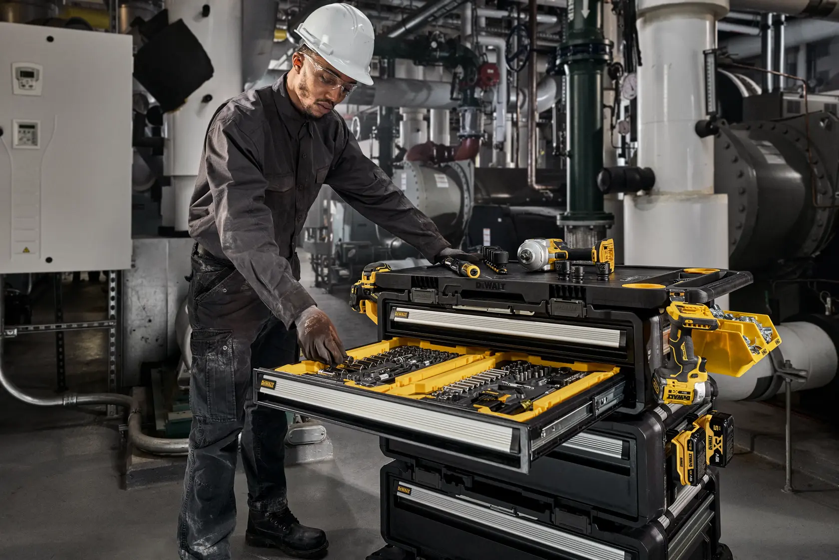 Une personne portant des vêtements de protection et un casque travaille avec un système de rangement d’outils DEWALT DWST60510-1 dans un environnement industriel. Le coffre à outils est ouvert et montre des outils et accessoires bien organisés dans plusieurs tiroirs.