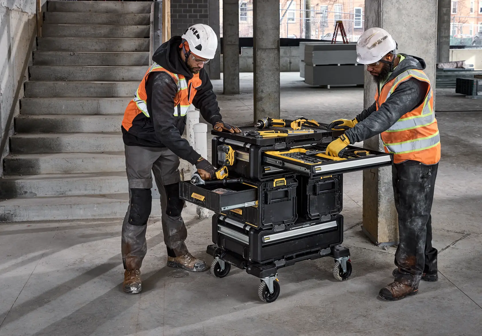 Deux ouvriers du bâtiment portant des casques de sécurité et des gilets réfléchissants utilisent un coffre à outils mobile DEWALT sur roulettes sur un chantier. Le coffre à outils possède plusieurs compartiments et tiroirs, et contient divers outils DEWALT. Les ouvriers accèdent activement aux outils, l'un ouvrant un tiroir et l'autre utilisant la surface de travail.
